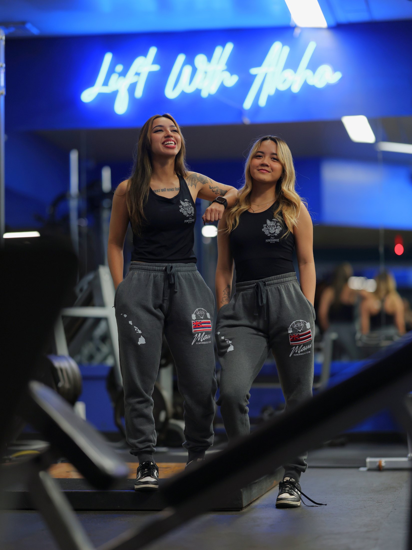 Two smiling Maui Powerhouse Gym members beneath the Lift With Aloha sign